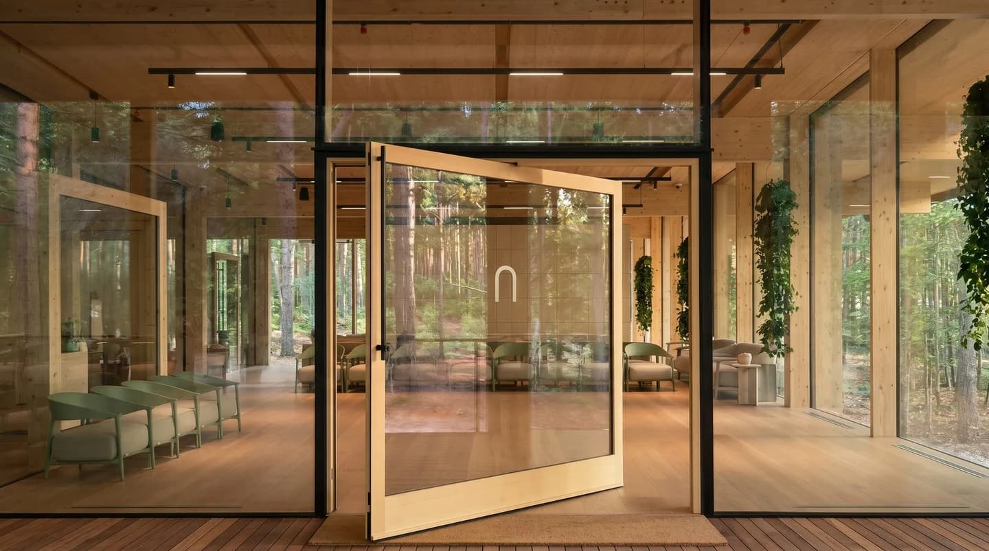 A glazed pivot door standing open into a timber lounge, forest beyond